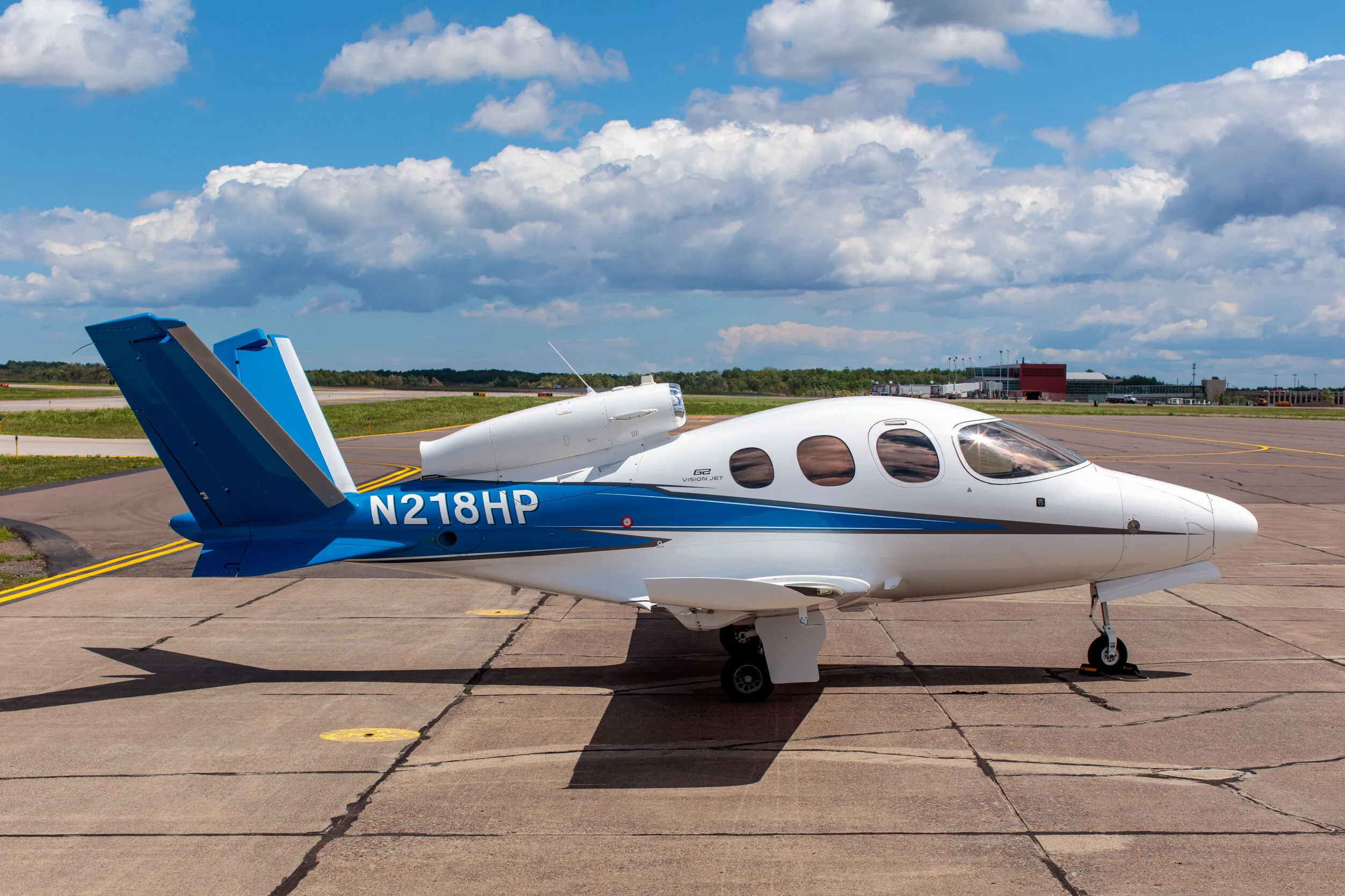 A white and blue Cirrus Vision Jet (N218HP) parked on a tarmac under a partly cloudy sky.