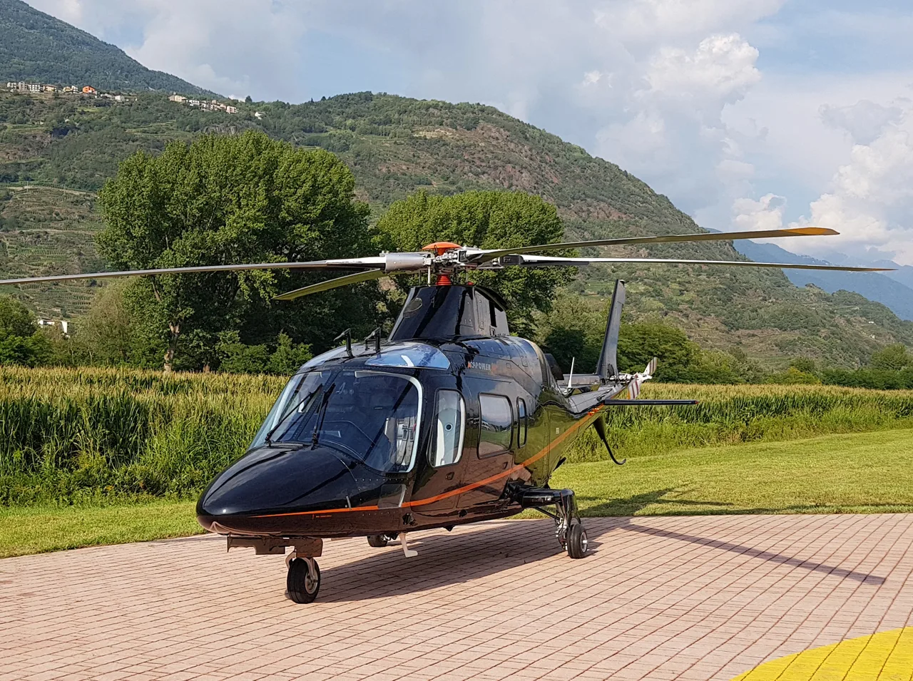 A black Agusta A109 Power helicopter with orange stripes is parked on a paved surface with green fields and mountains in the background.
