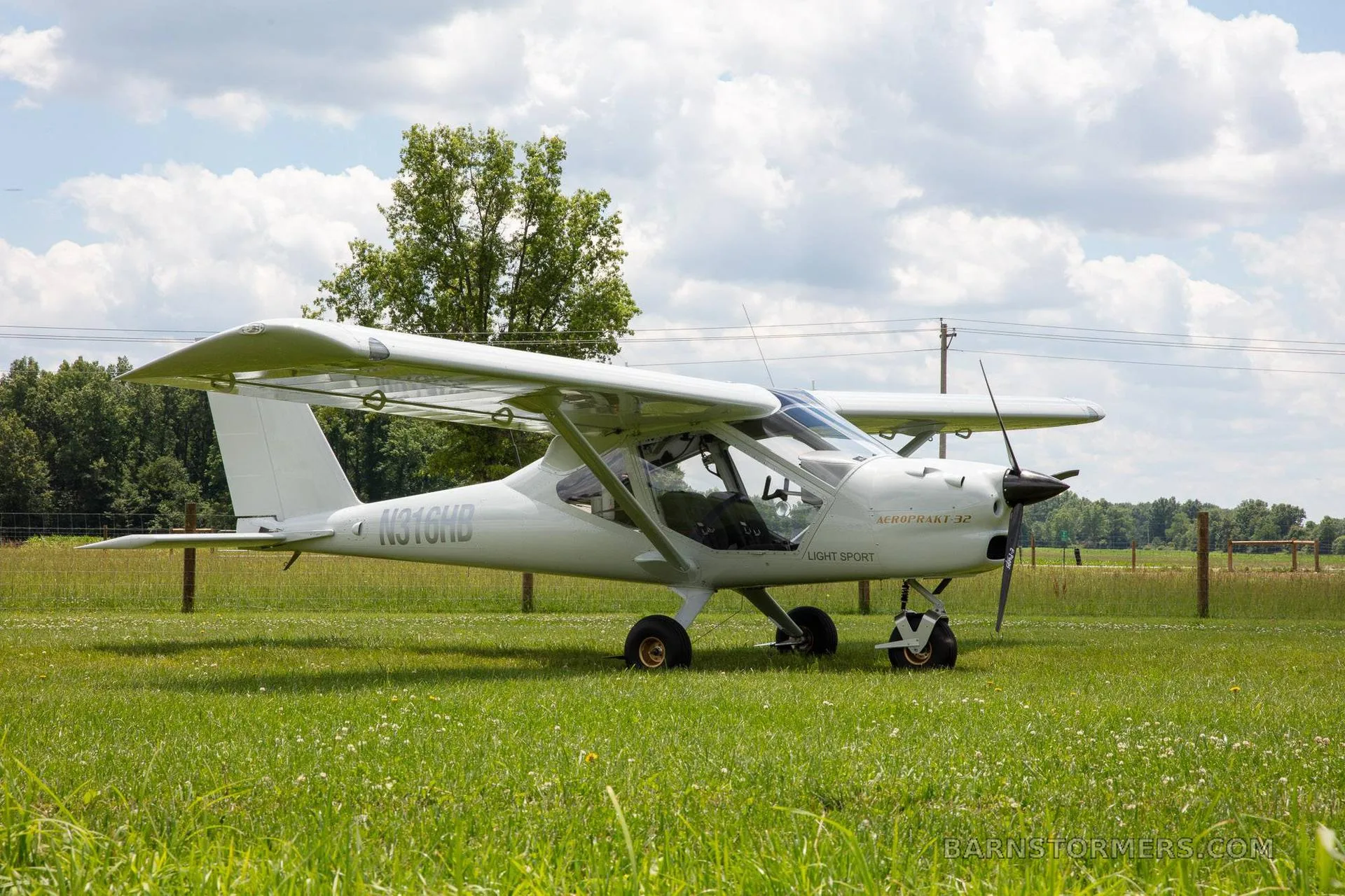 A white Aeroprakt-32 light sport aircraft with registration N316HB is parked on a green field with trees in the background.