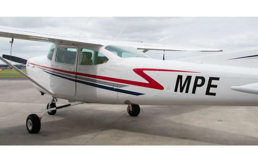A white single-engine propeller aircraft with red and blue stripes and tail number MPE, parked on an airfield.