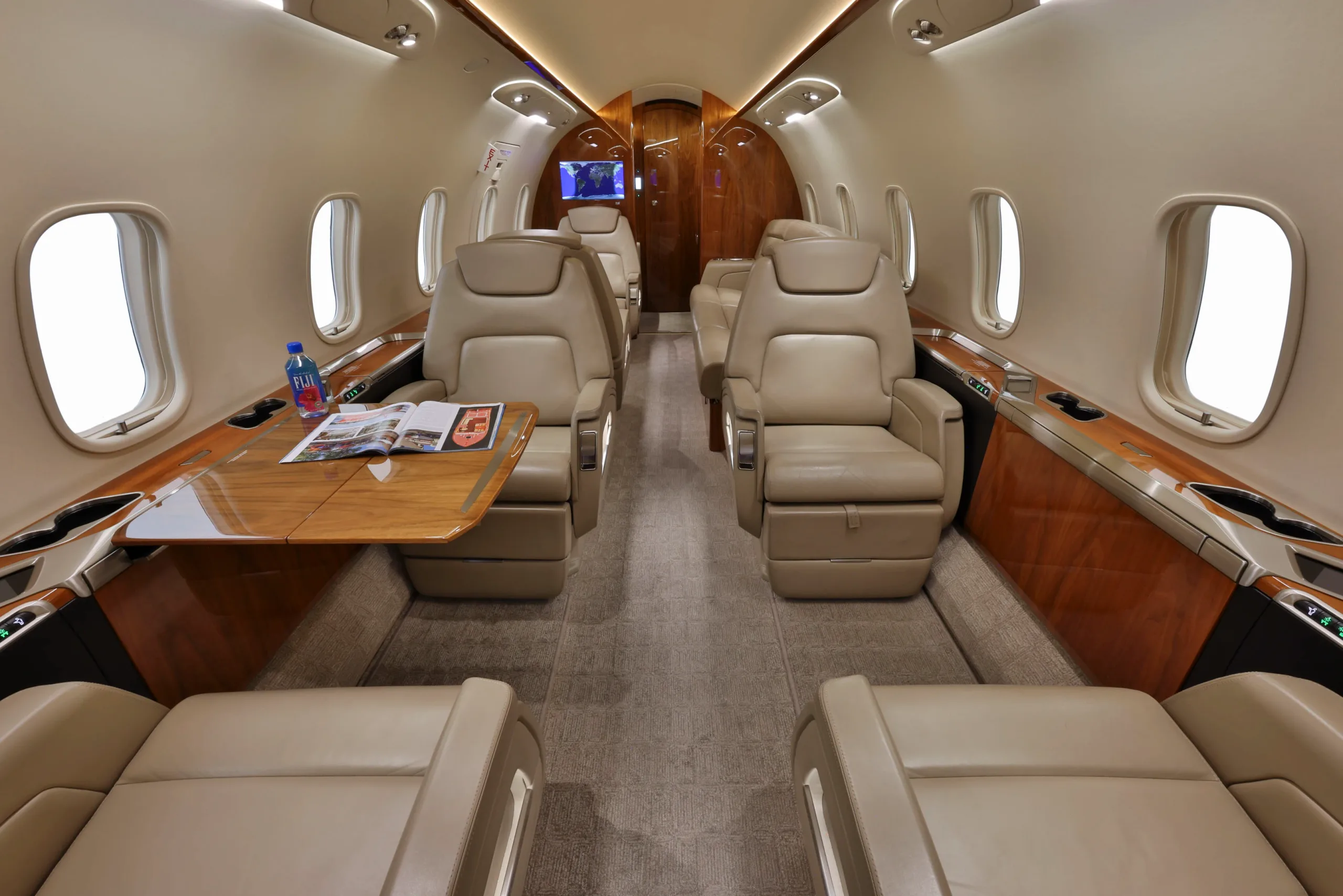The luxurious cabin interior of a business jet, featuring beige leather seats, a wooden table, and large windows.