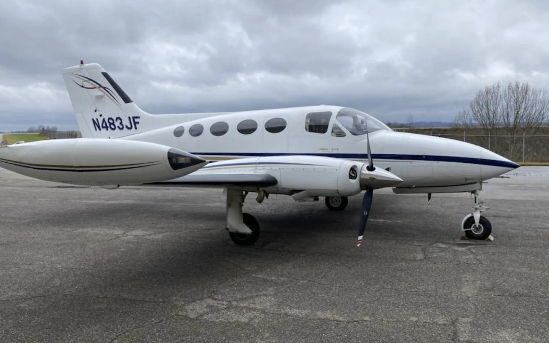 A white Cessna 414 Chancellor with blue and gold stripes, tail number N483JF, parked on an asphalt ramp under a cloudy sky.