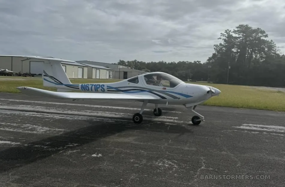 A white Diamond DA20-C1 Eclipse aircraft with blue and grey stripes, registration N671PS, parked on an asphalt surface next to a grassy area.
