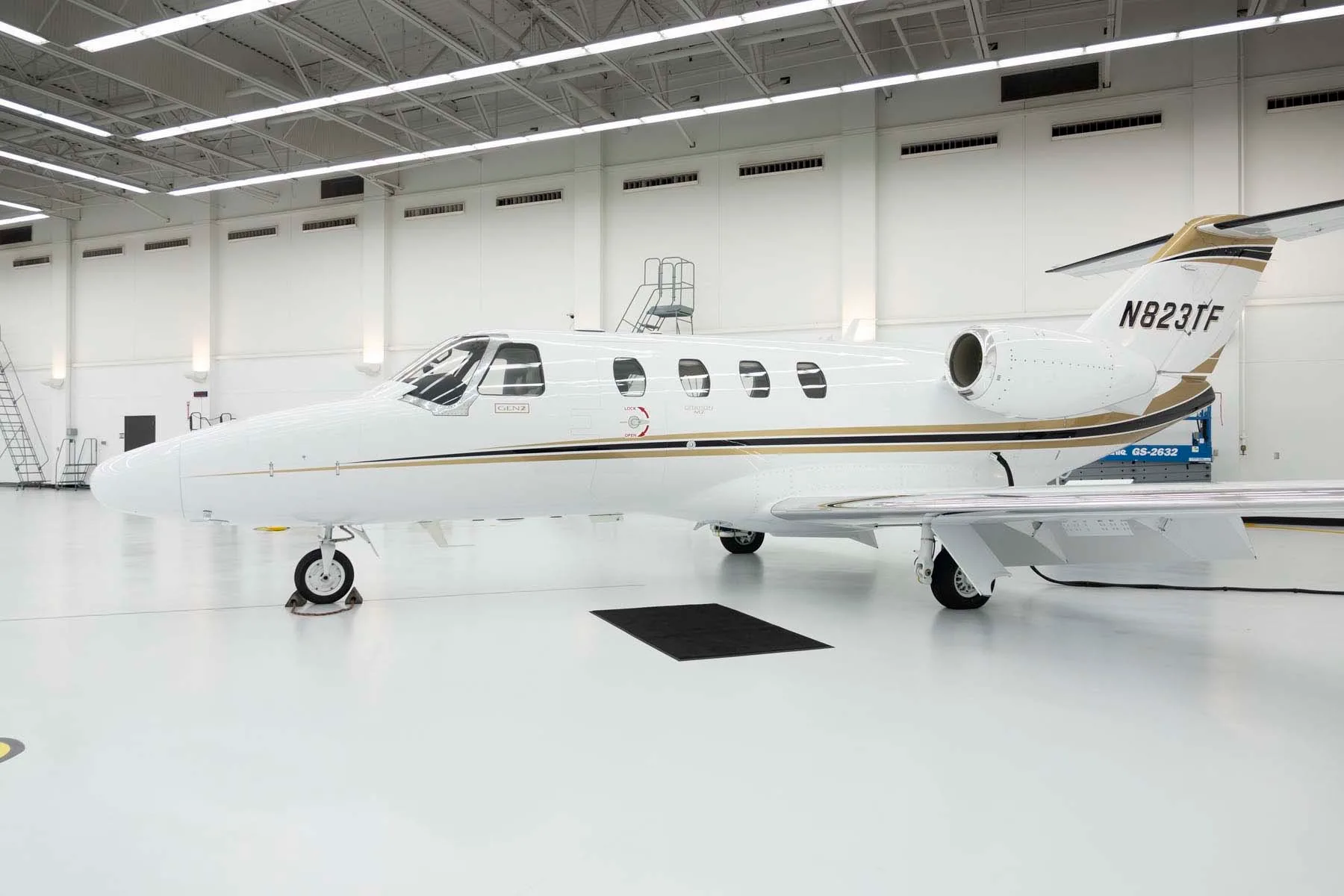 A three-quarter front view of a white Cessna Citation M2 jet with gold and black stripes, parked in a hangar.