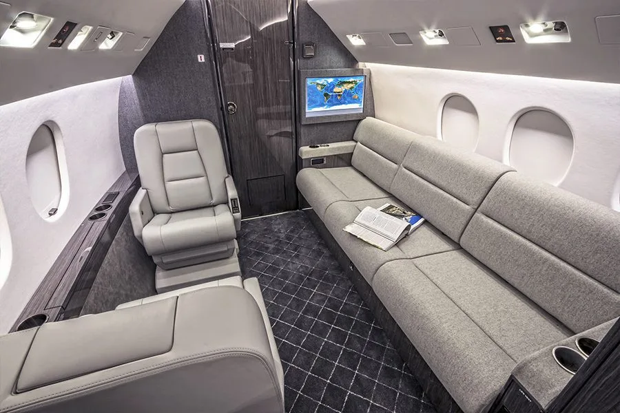A view of the private jet's cabin featuring a grey sofa, a single seat, and a wall-mounted map display.