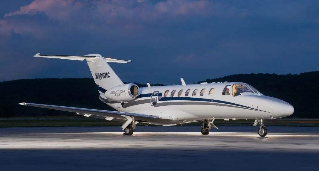 A white Cessna Citation CJ3 business jet (N906MC) with blue and grey stripes is parked on a tarmac at dusk.