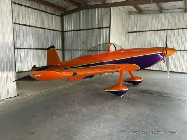 A side view of an orange and purple single-engine aerobatic aircraft parked in a hangar.