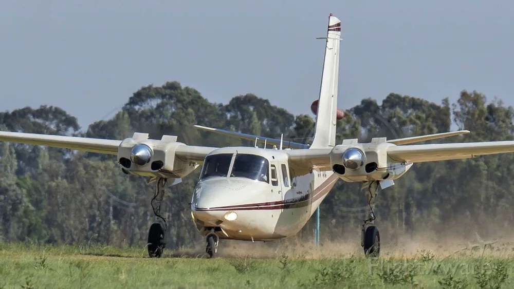 A white twin-engine aircraft with a maroon stripe on a grassy runway, viewed from the front-right.