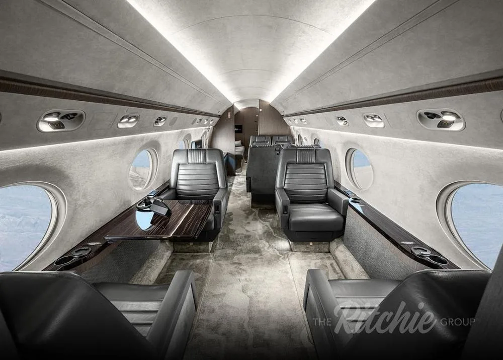 The luxurious interior cabin of a private jet features grey leather seats, a dark wood table, and large oval windows.