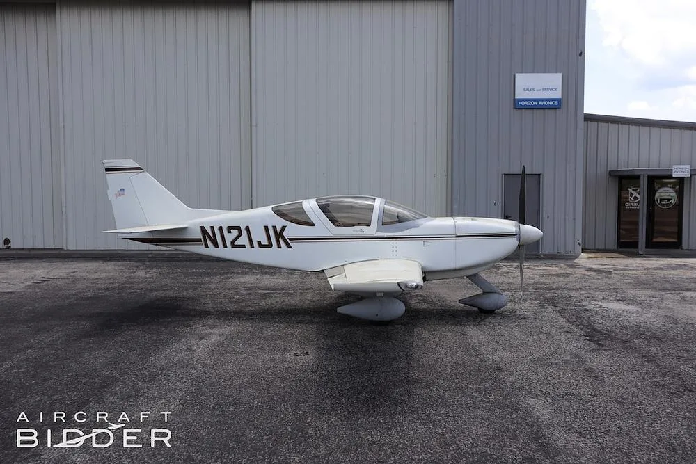 A side view of the white single-engine aircraft (N121JK) with brown stripes parked in front of a hangar.