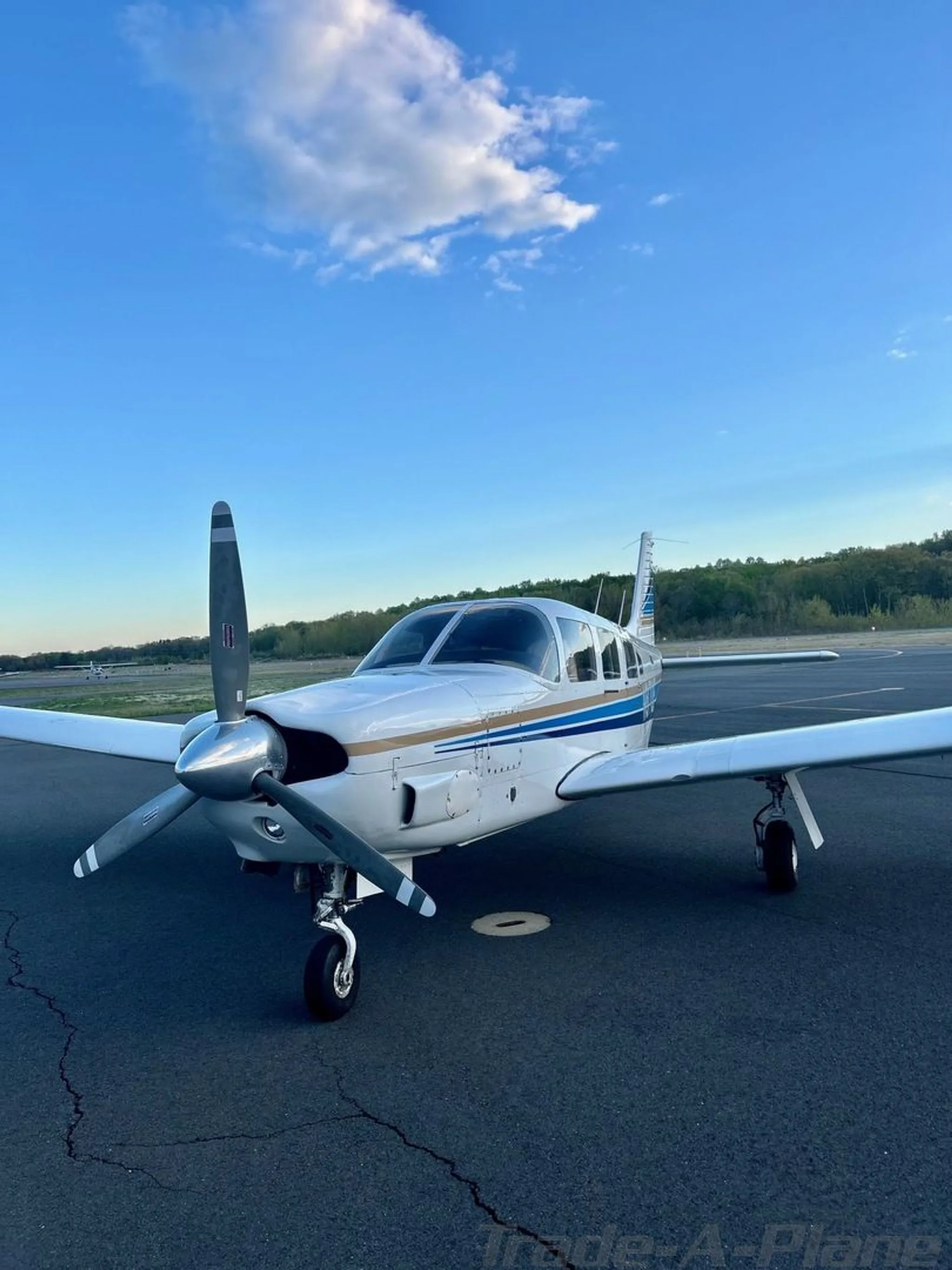 A front three-quarter view of a white Piper Cherokee aircraft with blue and gold stripes, parked on an airfield.