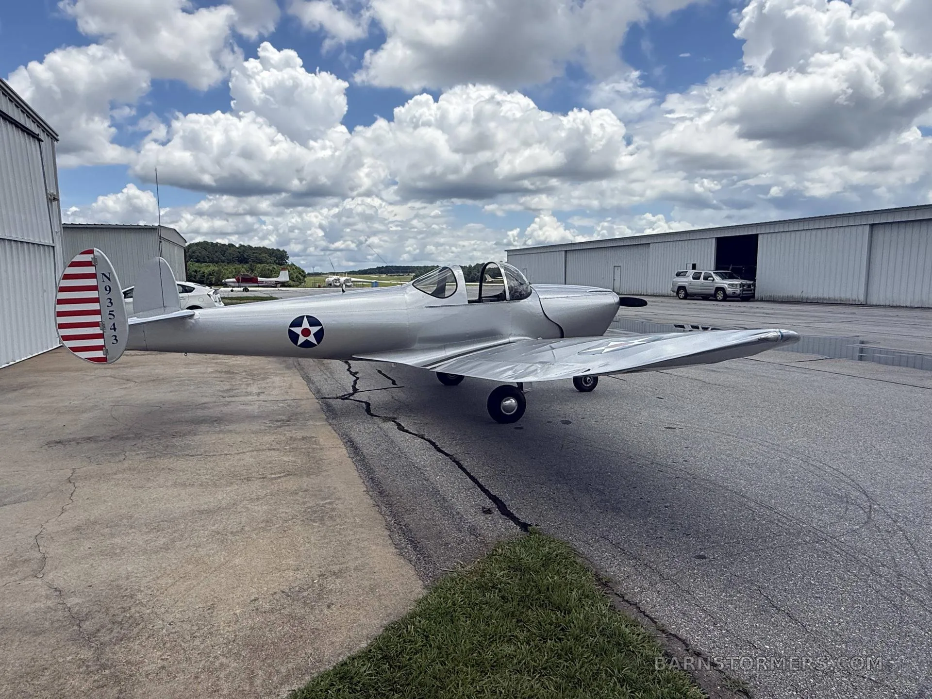 A silver single-engine aircraft with US military-style markings on the tail, parked on an airfield with hangars under a cloudy sky.