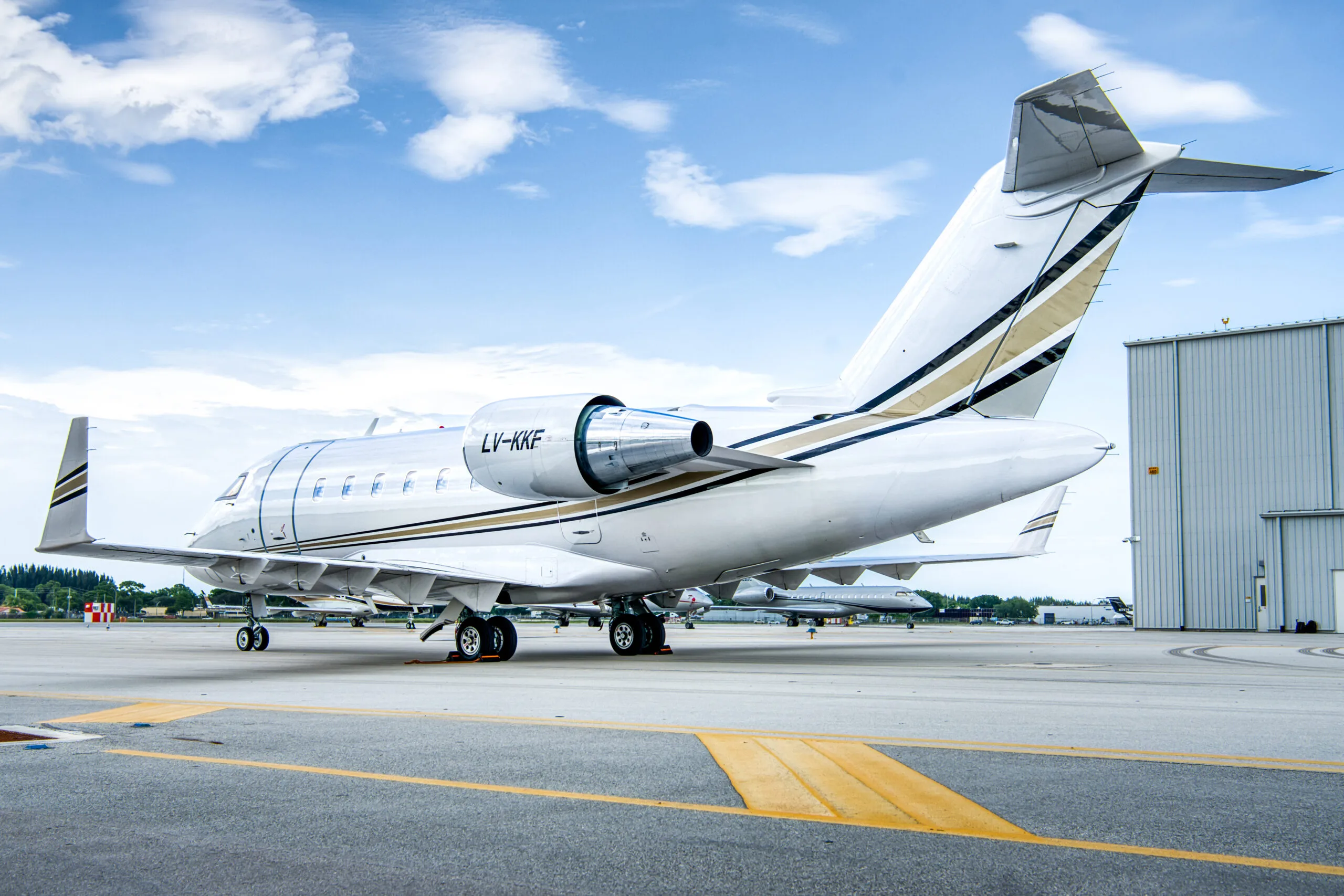 A white Bombardier Challenger business jet with gold and black stripes, registration LV-KKF, parked on an airport tarmac next to a hangar.