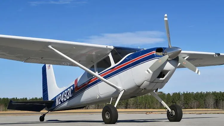 A white and blue Cessna 180 Skywagon with red stripes, registration N2493C, parked on an airfield under a clear blue sky.