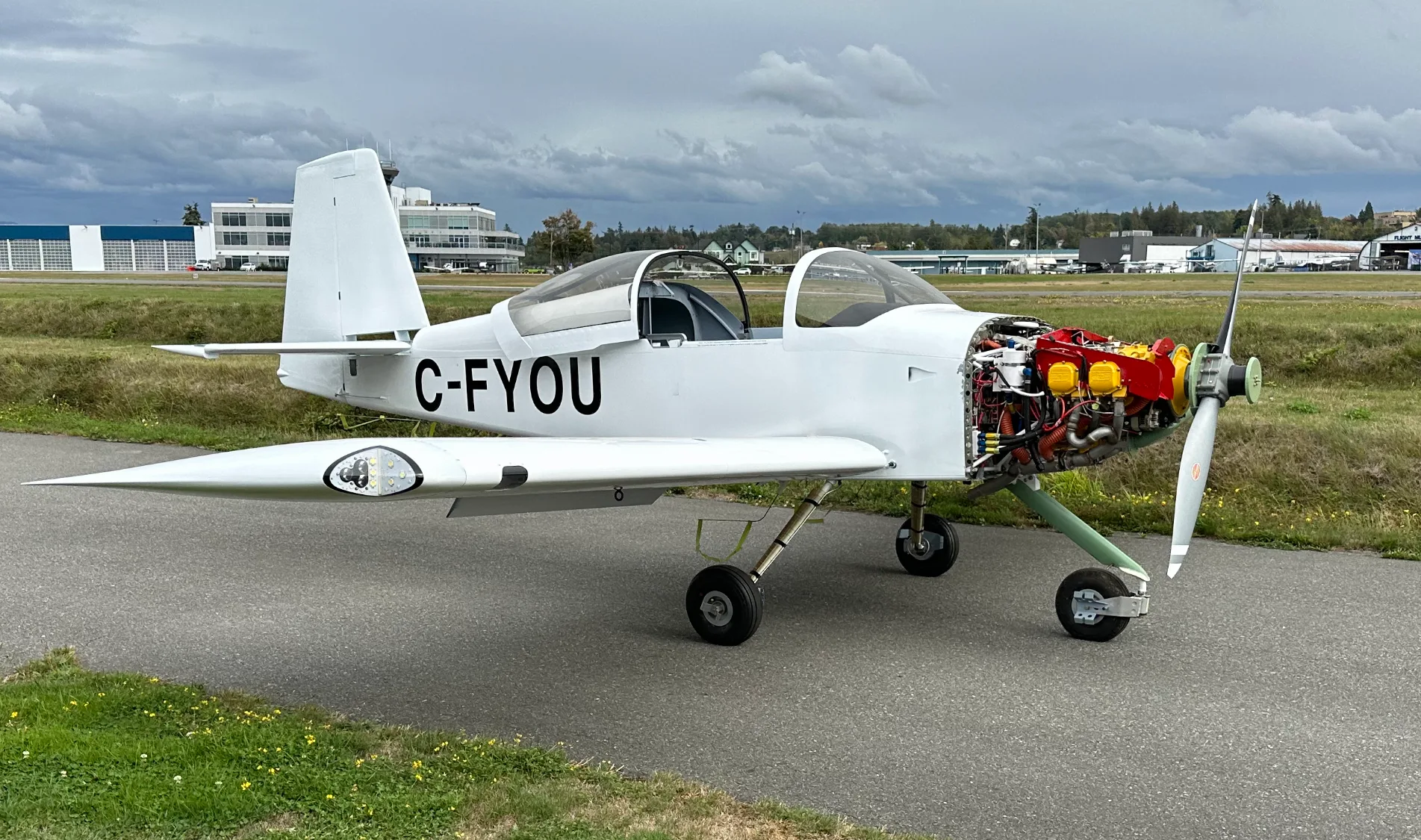 A white single-engine aircraft, registration C-FYOU, with its engine cowling removed, revealing the engine, parked on an asphalt apron at an airport.