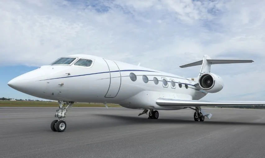 A white Gulfstream business jet with blue pinstripes is parked on an asphalt runway under a partly cloudy sky.