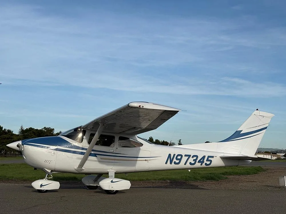 A white and blue Cessna aircraft with registration N97345 parked on an tarmac with trees and grass in the background under a partly cloudy sky.