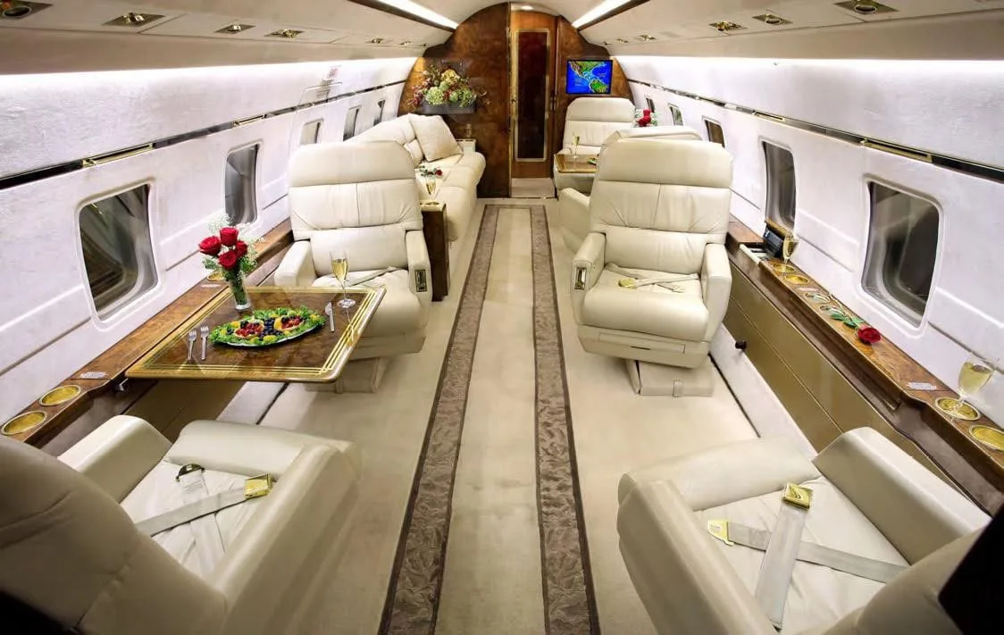 A luxurious aircraft cabin with cream leather seats, a dining table set with fruit, and wood accents.