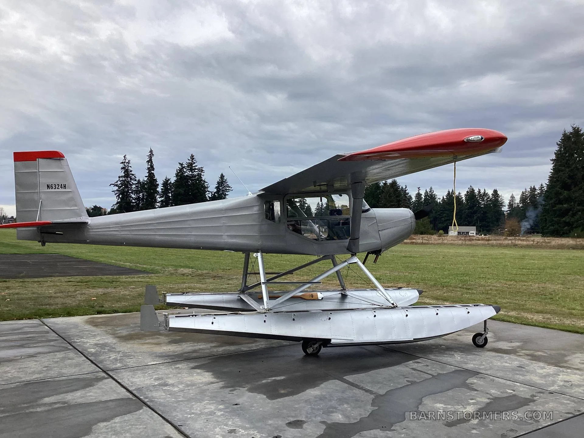 A silver floatplane with a red nose and tail, registration N6324H, parked on a concrete pad with a grassy field and trees in the background under a cloudy sky.