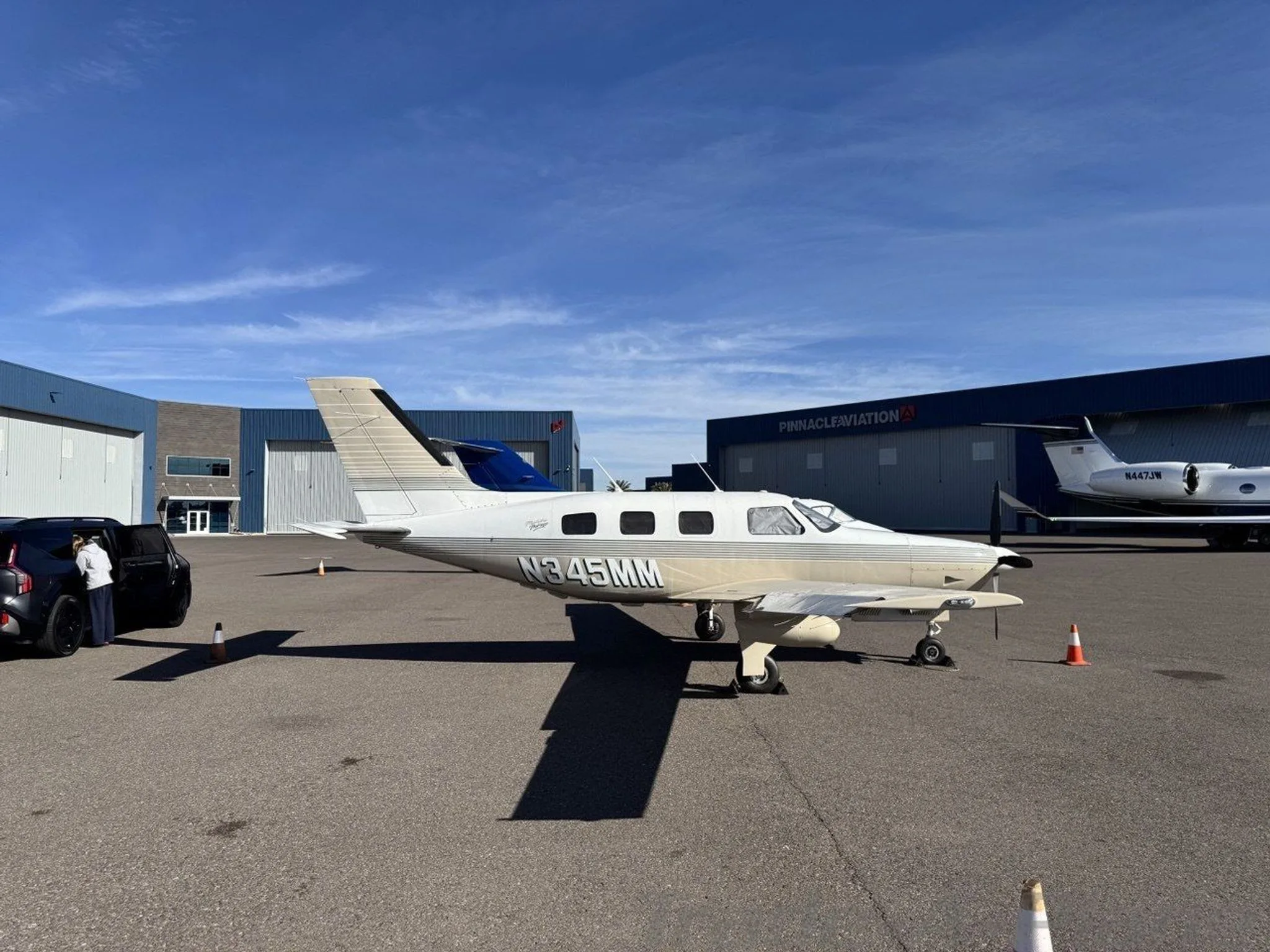 A white and tan single-engine propeller aircraft (N345MM) parked on an airfield with hangars in the background.