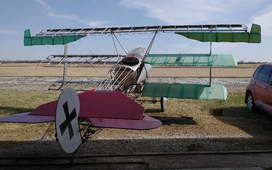 A triplane frame with silver fuselage, green wings, and a red tail with German cross insignia, parked on a grassy field.