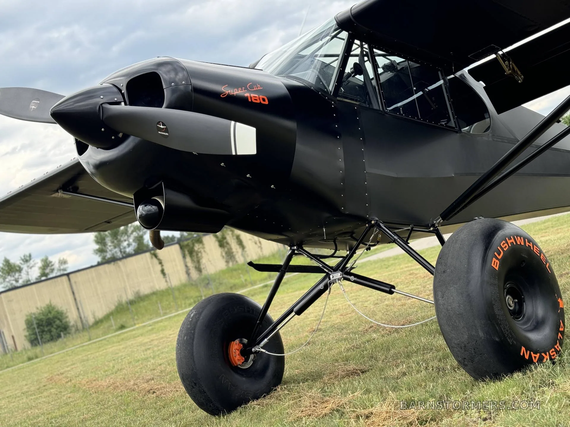 A black Super Cub 180 aircraft with large bush wheels, viewed from the front-right on a grassy field.