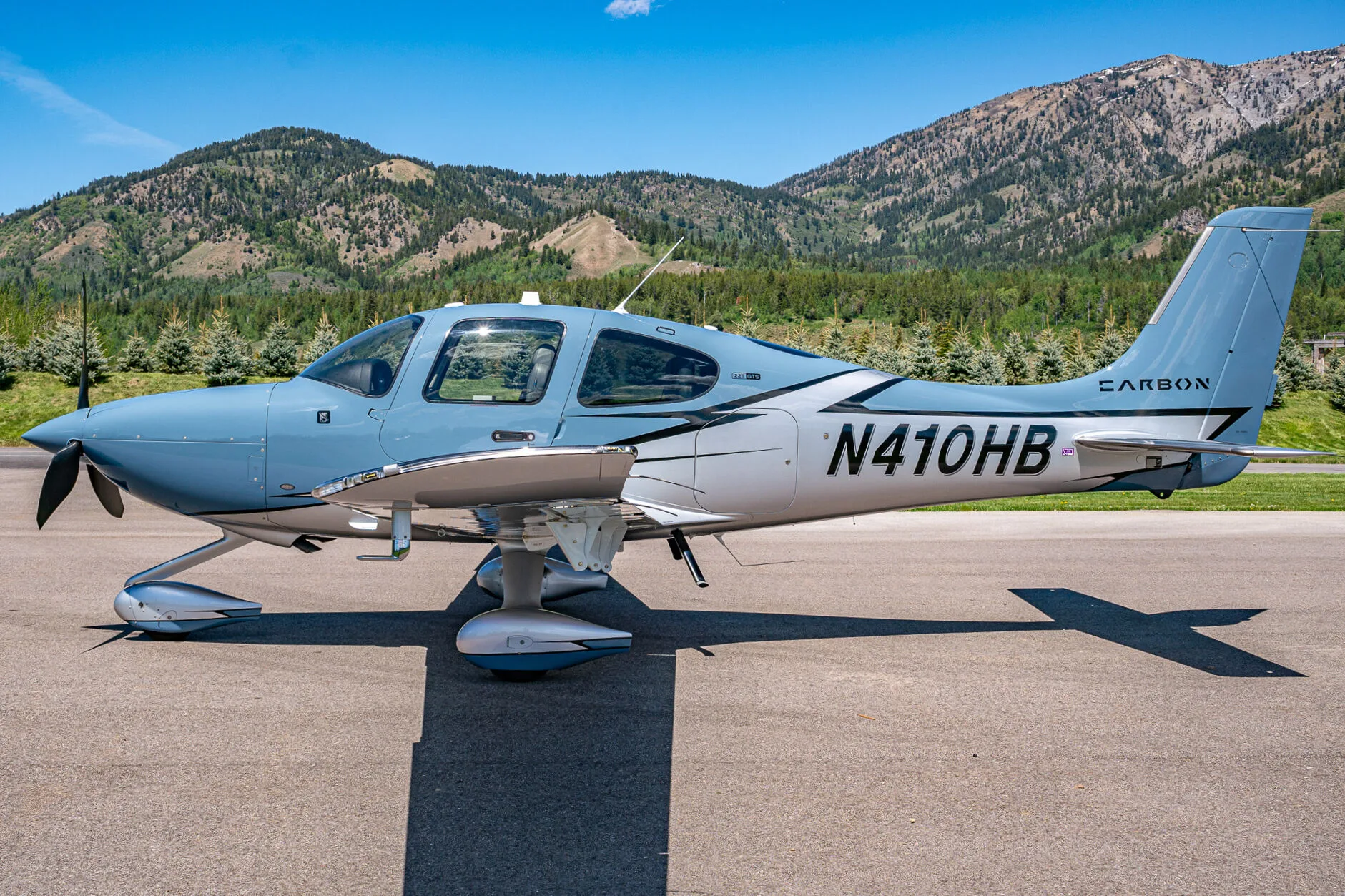 A light blue and silver Cirrus SR22T aircraft (N410HB) with 'CARBON' livery parked on an airfield with mountains in the background.