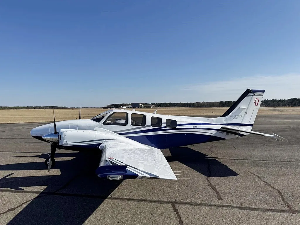 A white and blue Beechcraft Baron 55 twin-engine aircraft is parked on an airport tarmac under a clear sky.