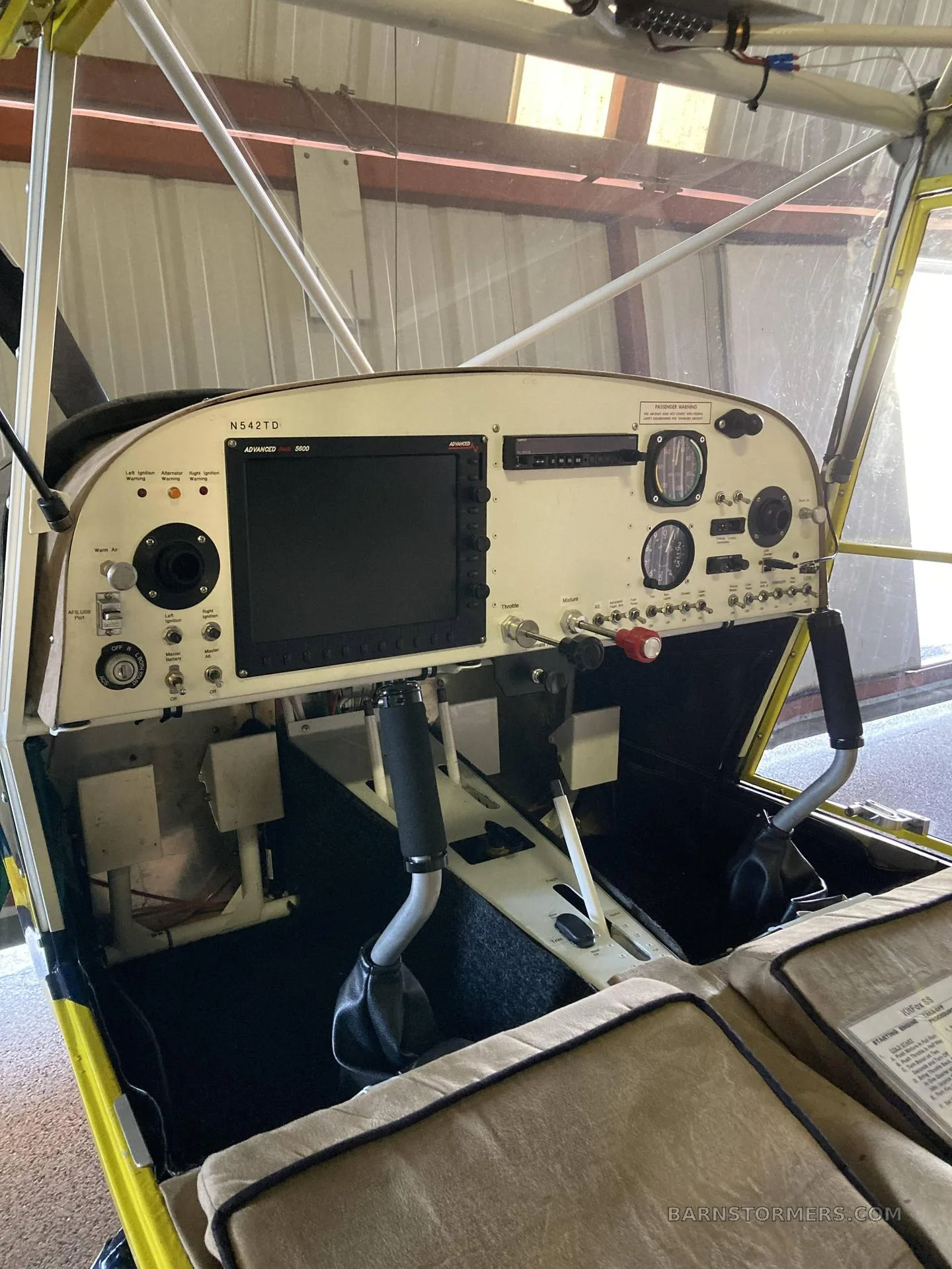 The cockpit of a blue and yellow aircraft, showing the instrument panel, dual controls, and tan fabric seats.