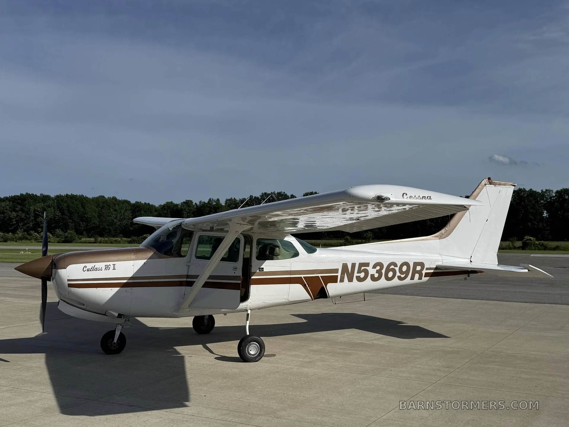 A white and brown Cessna Cutlass RG II (N5369R) is parked on an asphalt ramp under a partly cloudy sky.