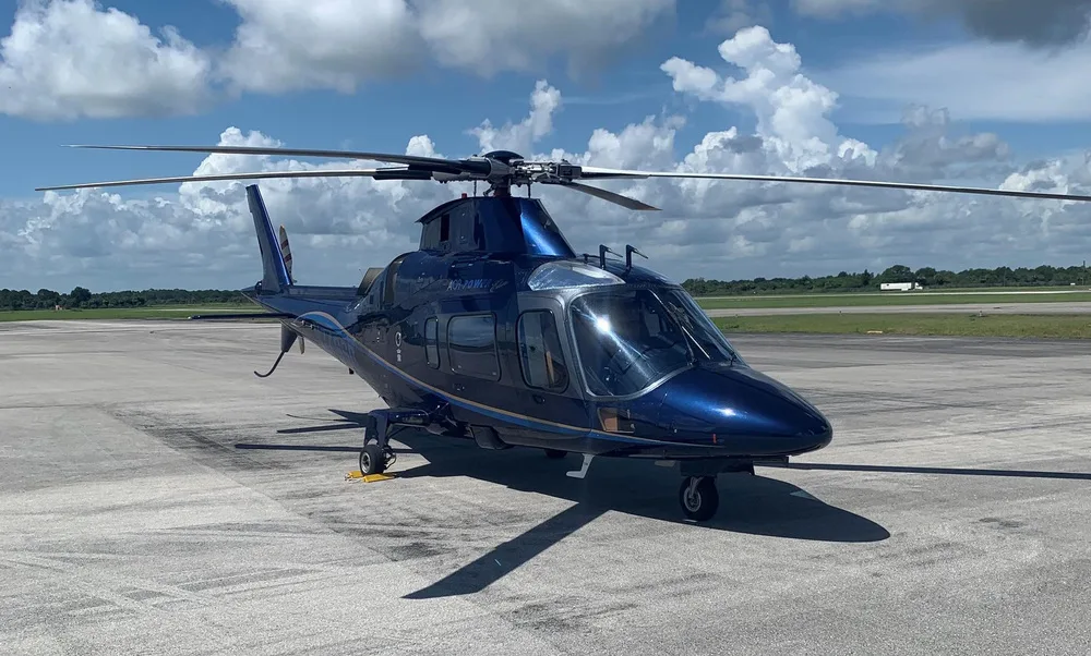 A dark blue Leonardo AW109 helicopter with gold stripes sits on an airfield under a partly cloudy sky.