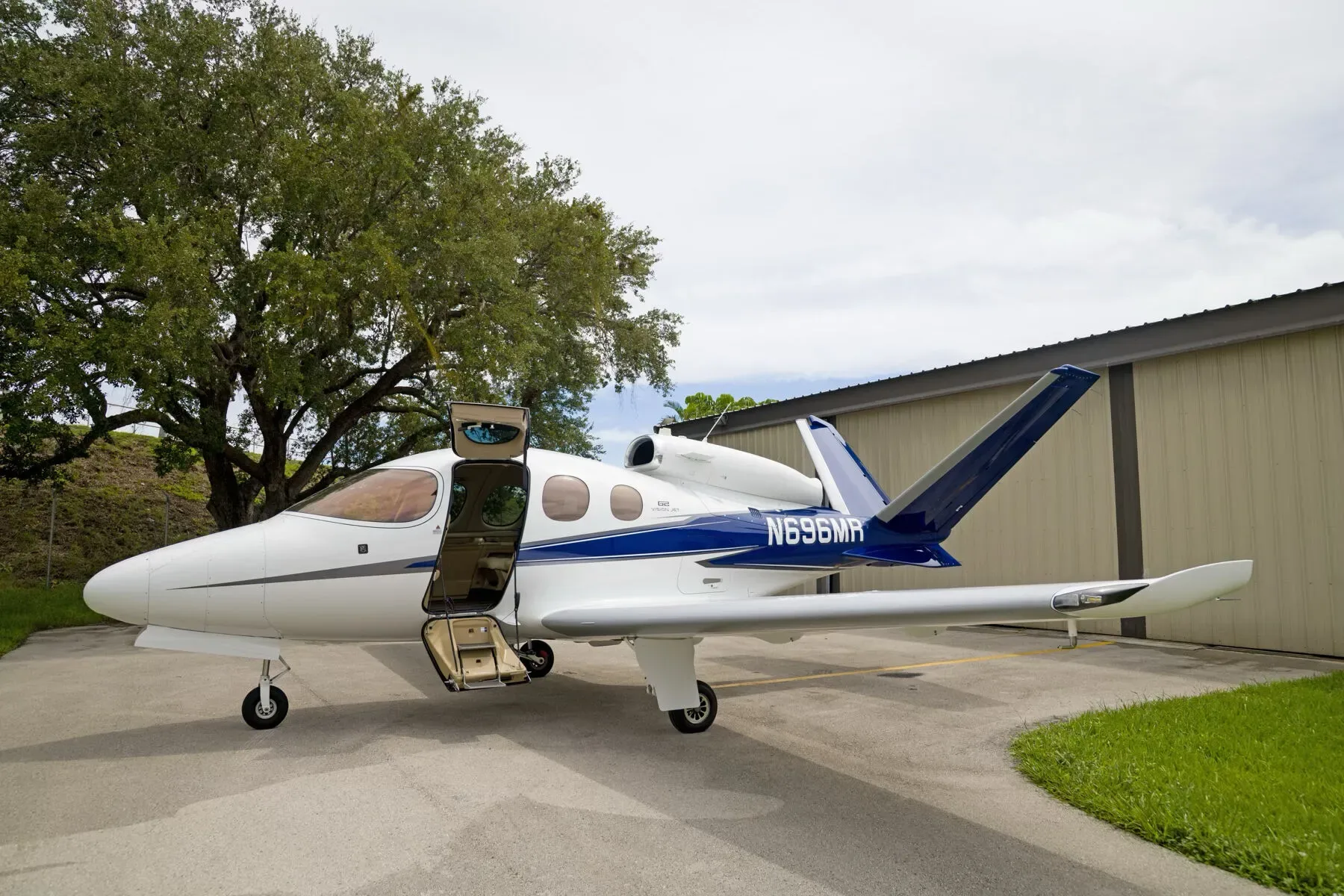 A white Cirrus Vision Jet (N696MR) with blue and grey stripes, parked on a tarmac with its main cabin door open.