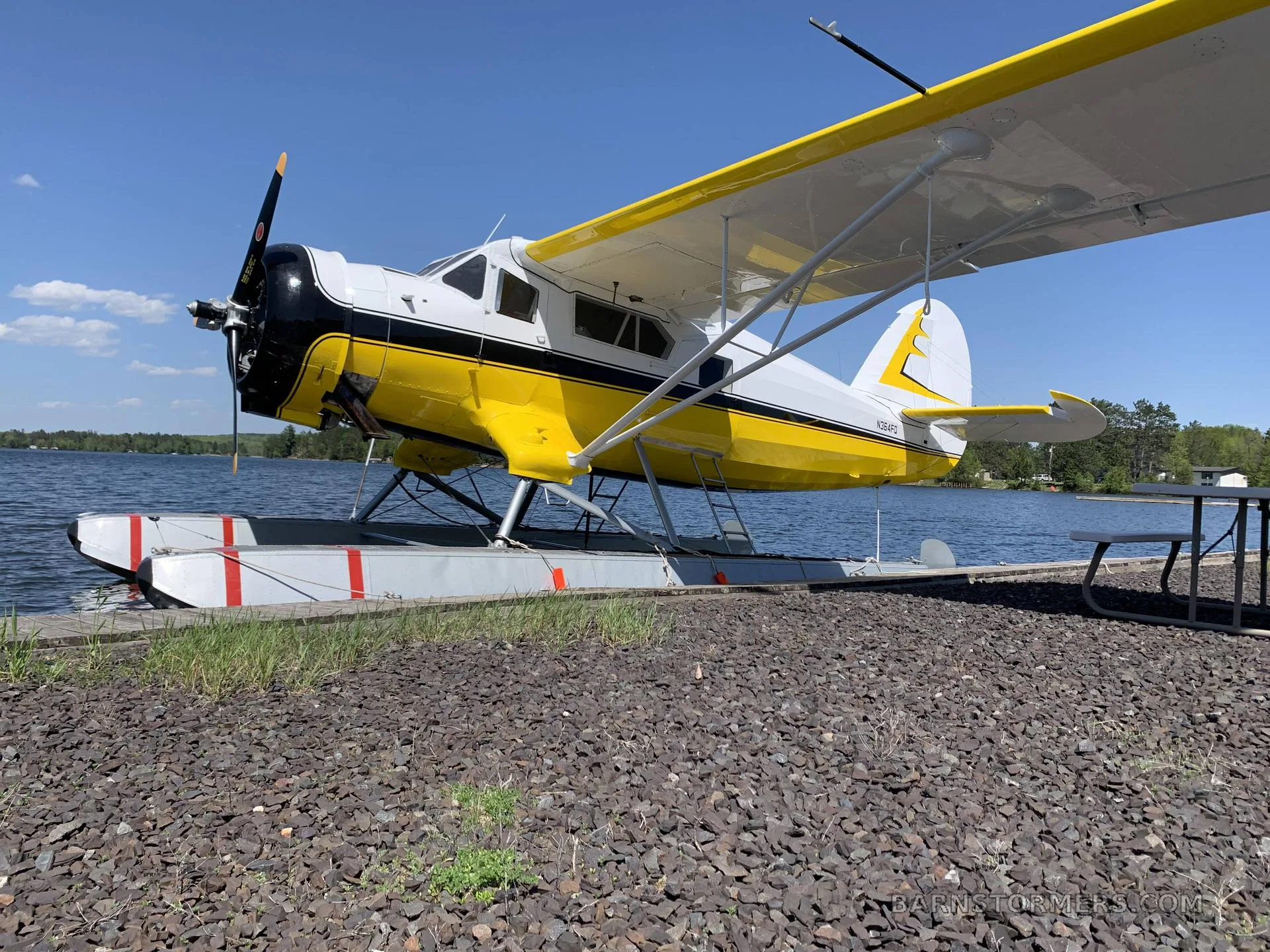 A yellow and white floatplane is docked at a rocky shore on a sunny day, with a lake and trees in the background.