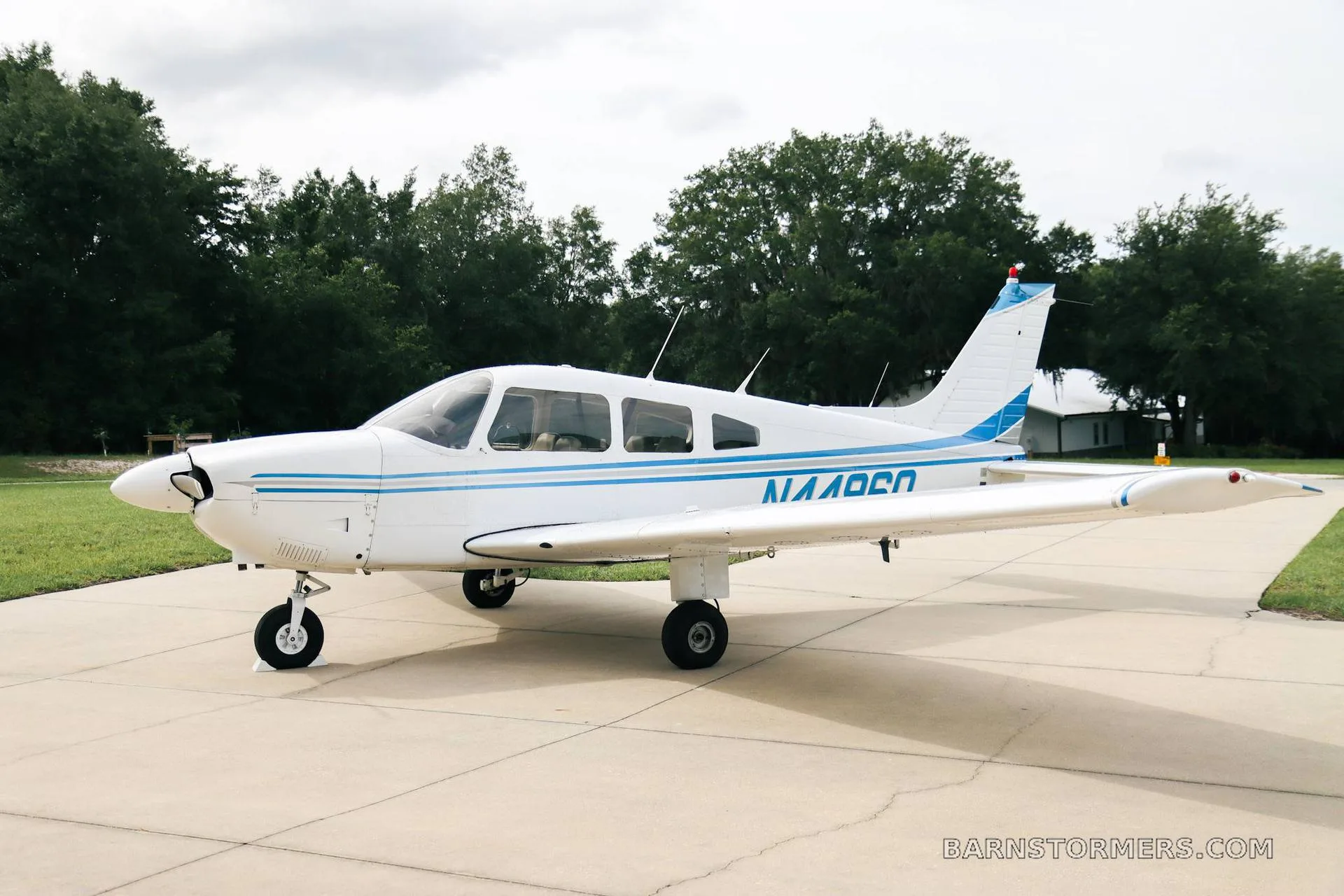 A white Piper PA-28 series aircraft with blue stripes, viewed from the front-right, parked on a concrete pad.