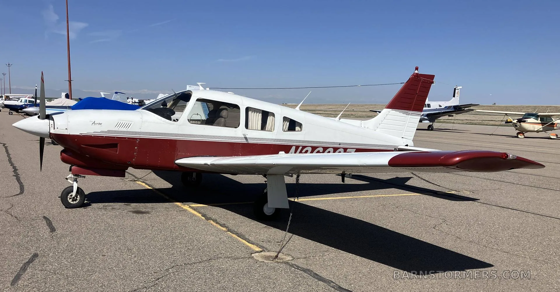 A white and maroon Piper Arrow single-engine aircraft parked on an airport tarmac under a clear blue sky.