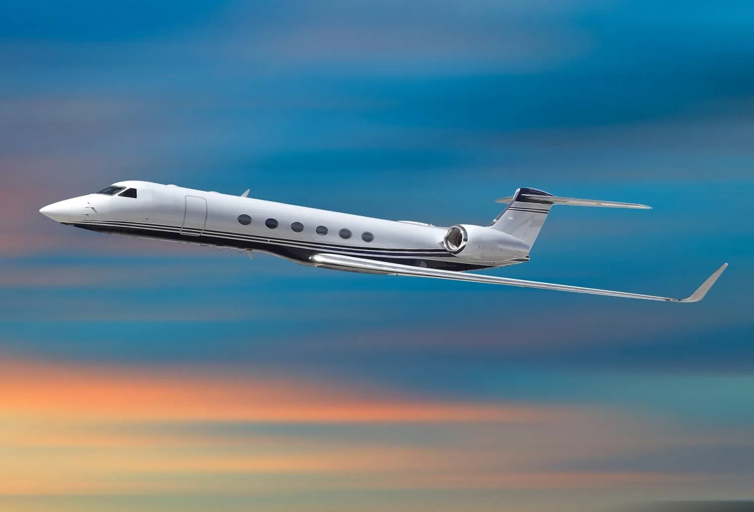 A white Gulfstream-style business jet with a black stripe flying against a vibrant blue and orange sky.
