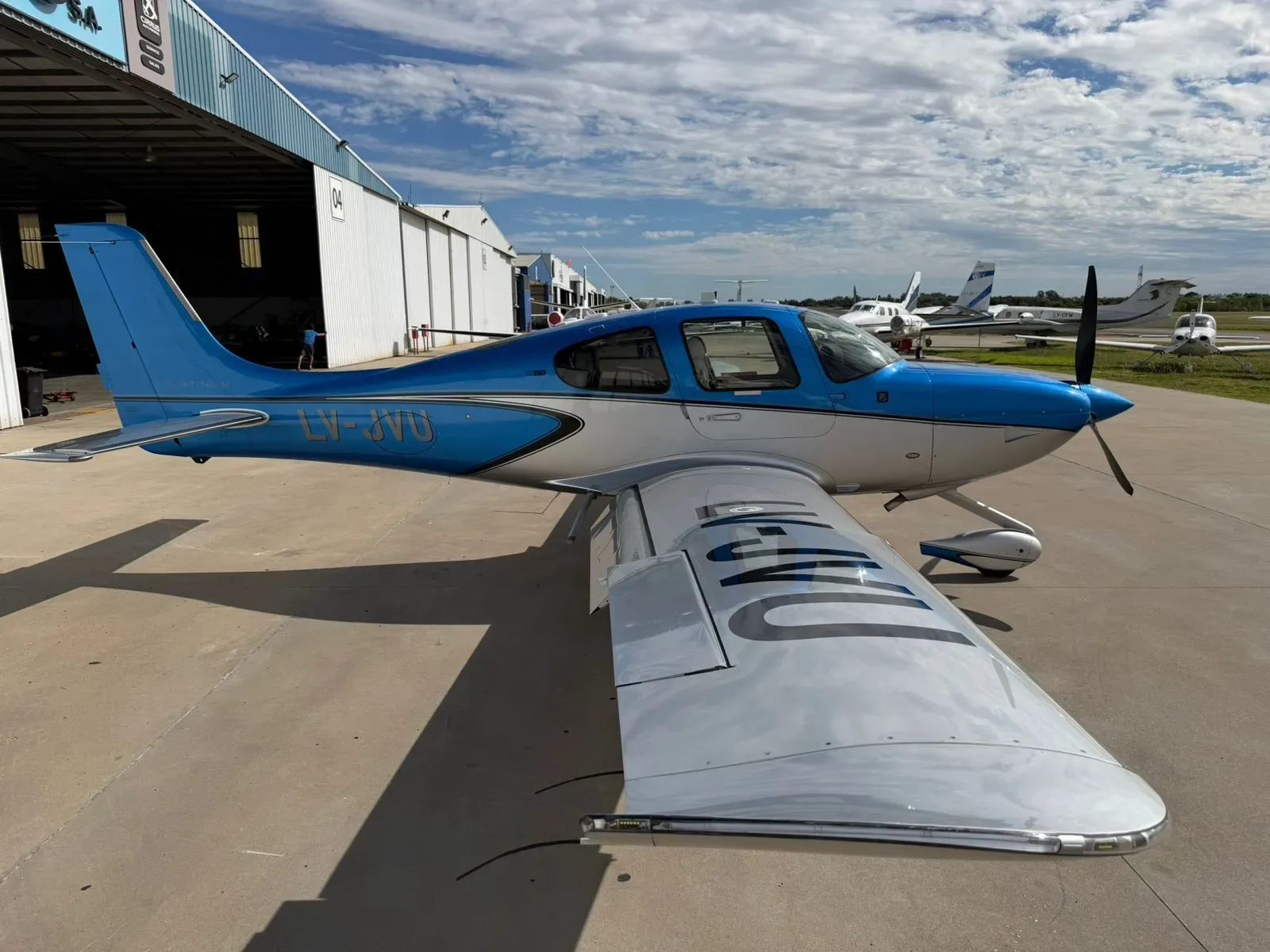 A blue and silver Cirrus SR22 aircraft, registration LV-JYU, parked on an airfield with a hangar in the background.