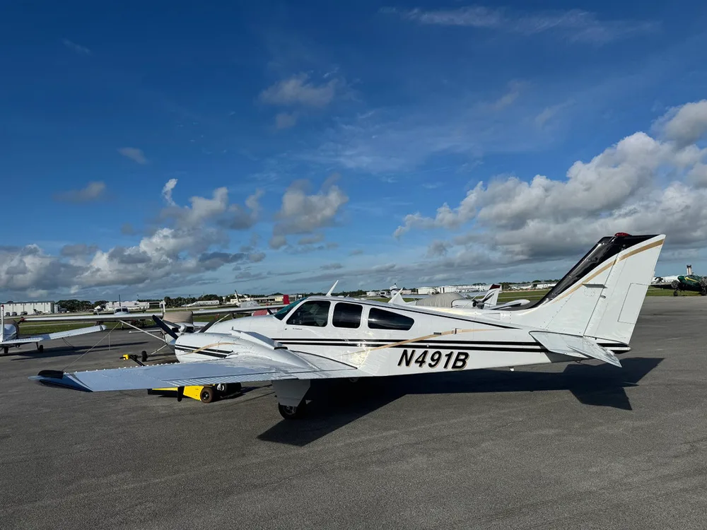 A white twin-engine Beechcraft Baron aircraft with black and gold stripes, registration N491B, parked on an airfield under a blue sky.