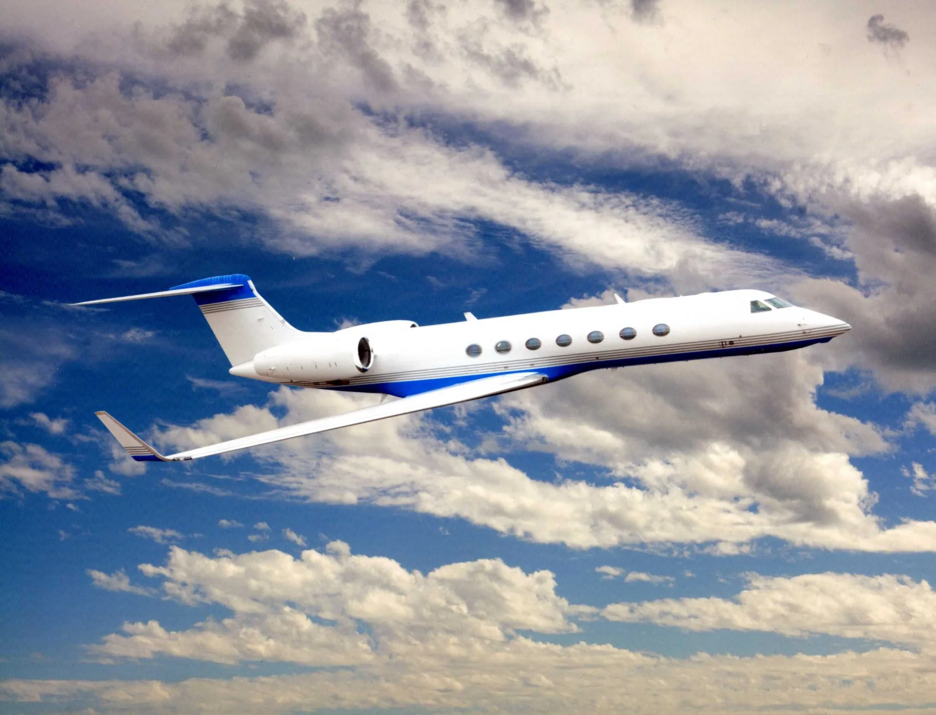 A white business jet with blue stripes flies through a partly cloudy blue sky.