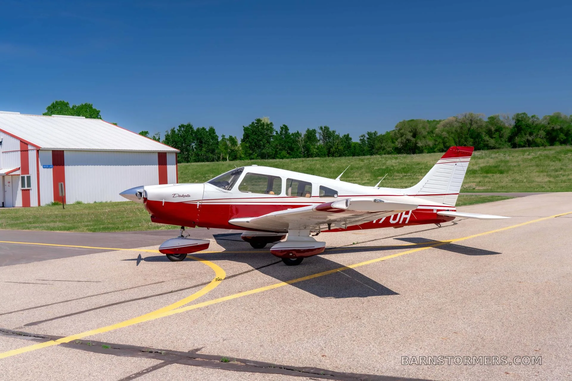 A red and white Piper Dakota aircraft is parked on an asphalt tarmac next to a white hangar.