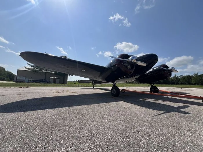 A dark twin-engine vintage aircraft parked on an asphalt runway under a sunny sky.