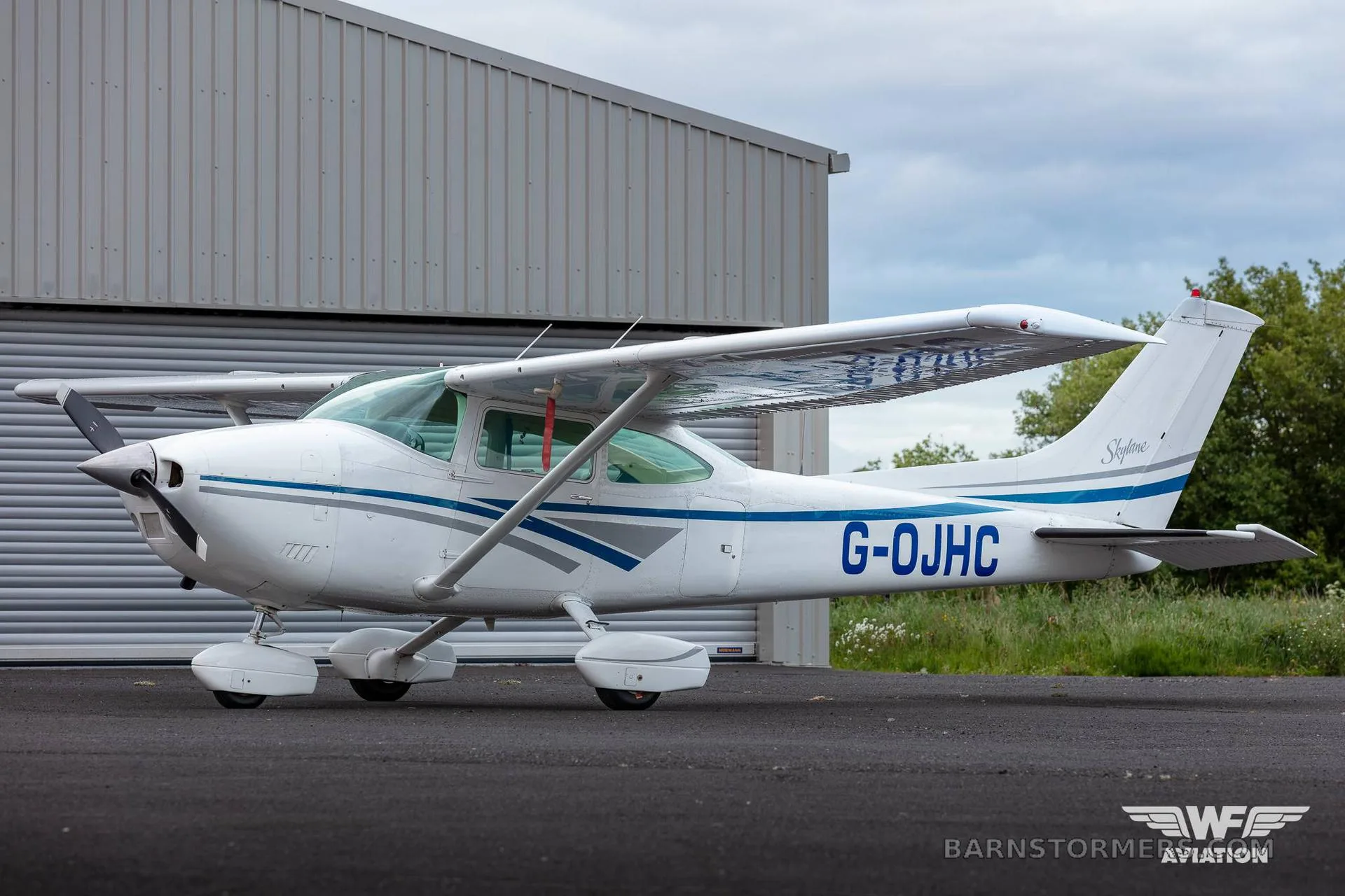 A white Cessna 182 Skylane with blue and grey stripes is parked on an tarmac in front of a hangar.