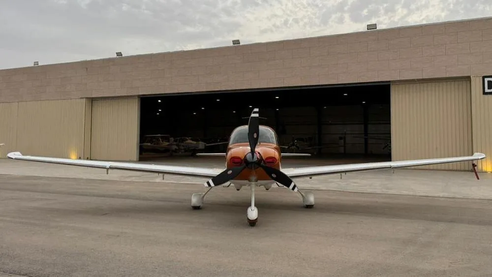 A front view of an orange and white Cirrus SR22 aircraft parked outside a hangar.