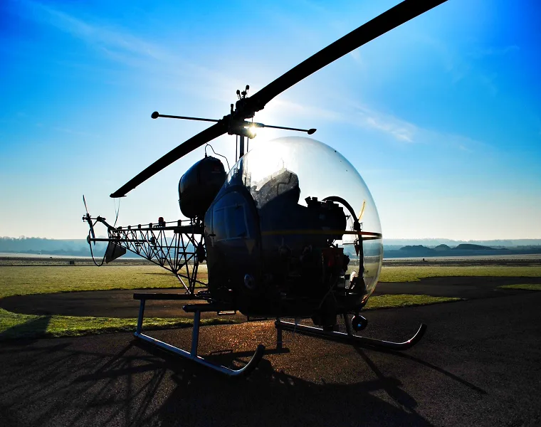 A silhouette of a bubble-canopy helicopter, possibly a Bell 47, parked on an airfield at sunrise or sunset.