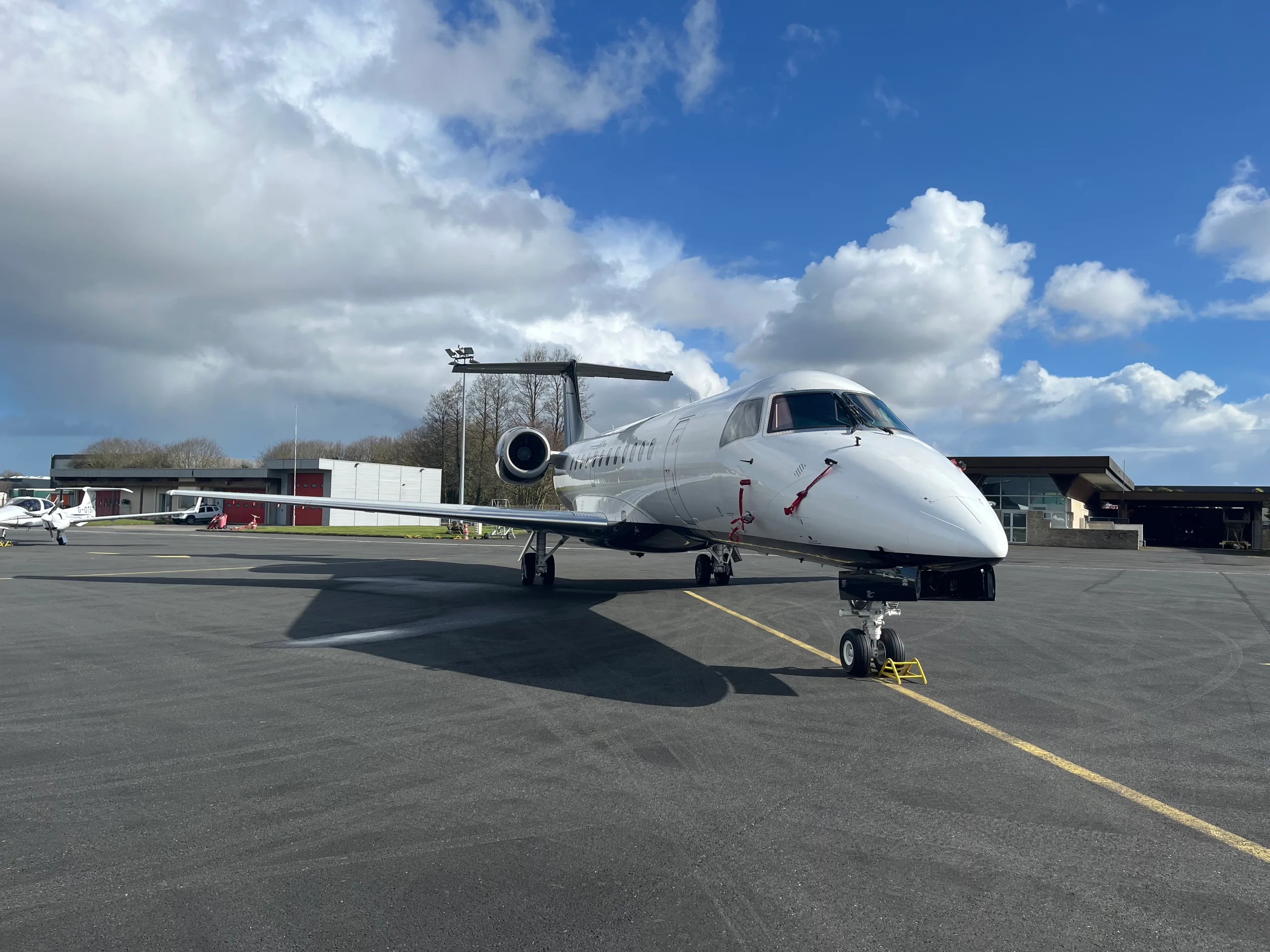 A white business jet with dark engines and tail, parked on an asphalt tarmac under a bright blue sky with white clouds.