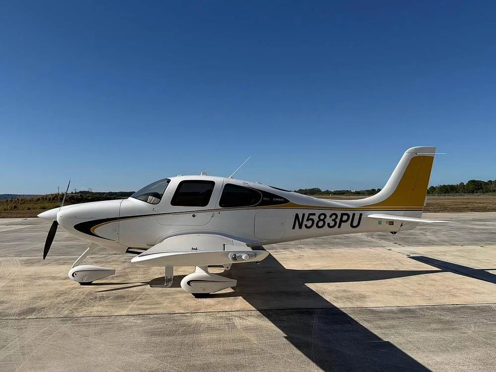 A white Cirrus SR22 aircraft with black and gold stripes and a gold tail, registration N583PU, parked on an airfield under a clear blue sky.