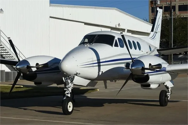 A white twin-engine turboprop aircraft with blue and grey stripes is parked on an apron in front of a white hangar.