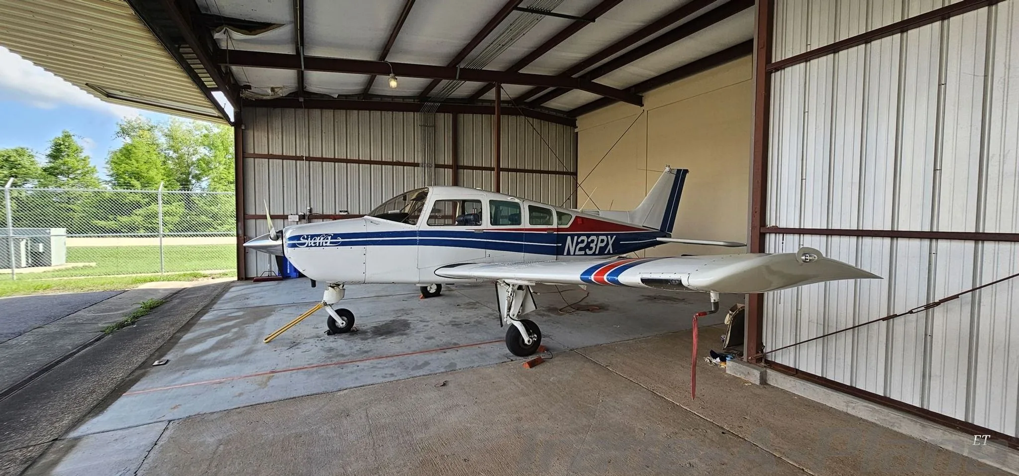 A white Beechcraft Sierra N23PX with blue and red stripes parked in a hangar, viewed from the front left.