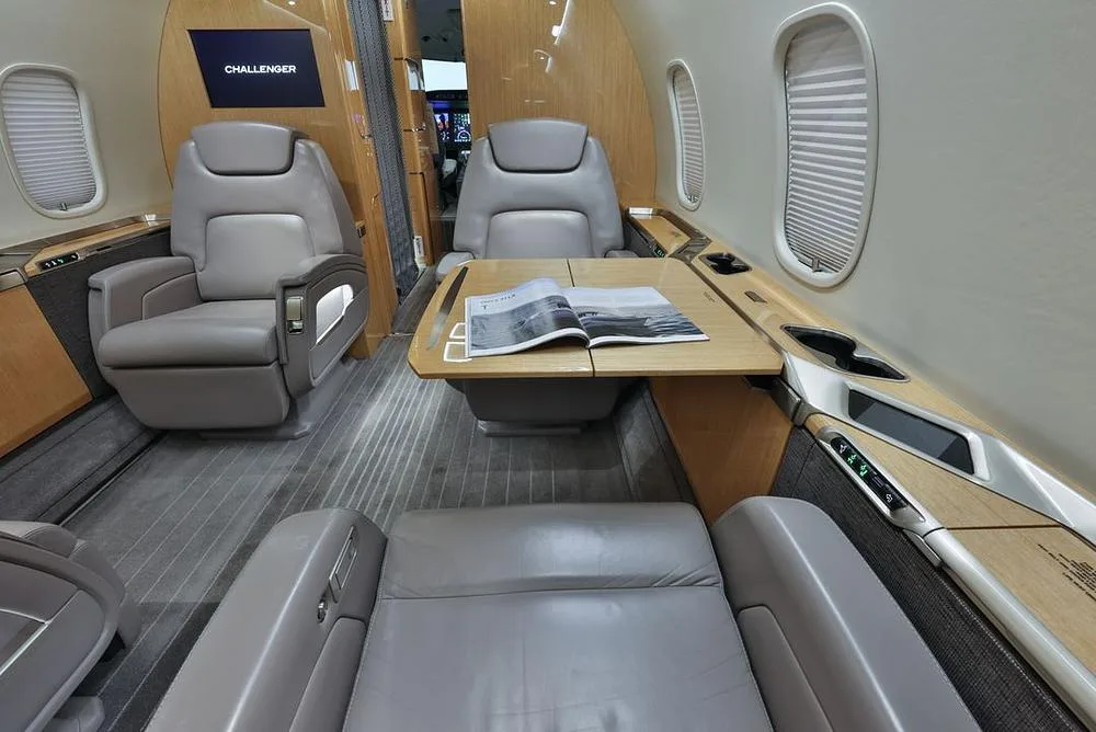 A view inside the business jet cabin, showing two grey leather seats facing a wooden table with a magazine, and a "CHALLENGER" screen in the background.