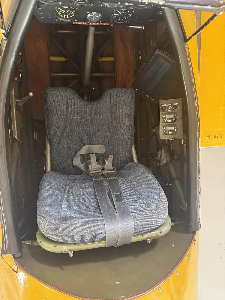 The rear cockpit of a yellow Stearman biplane, featuring a blue fabric seat with a harness.